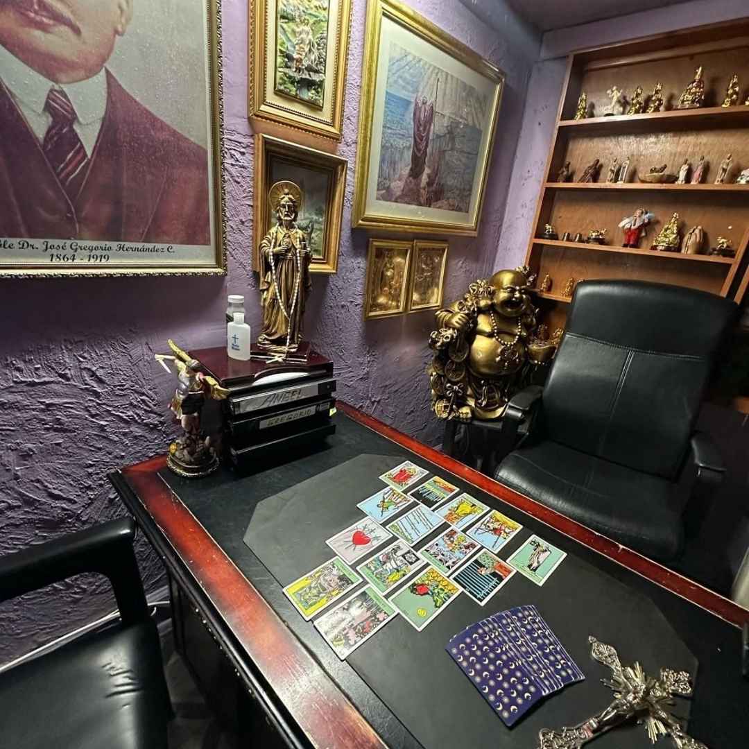 Mesa de lectura de tarot con cartas desplegadas, crucifijo, estatuas de santos y Buda en un consultorio espiritual esotérico.
