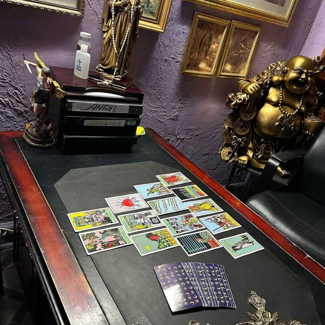 Mesa de consulta esotérica de Curandero Blanco con lectura de cartas del tarot, crucifijo, figuras religiosas y estatua dorada de Buda.