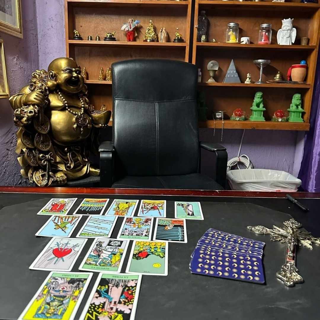 Mesa de lectura de tarot con cartas desplegadas en patron, crucifijo plateado y estatua dorada del Buda de la abundancia en consultorio esoterico.